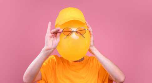 Osoba ubrana w żółty, luźny t-shirt i żółtą czapkę z daszkiem. Twarz ma zasłoniętą żółtym balonem, a na nim trzyma prawą ręką okulary w okolicach oczu. Zdjęcie na różowym tle.