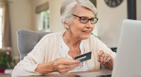 Kobieta w wieku 60+ robi zakupy online, w jednej ręce trzyma kartę kredytową.